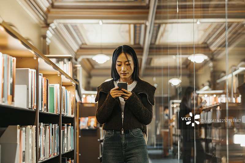 一名成年女学生在大学图书馆里站着使用智能手机图片素材