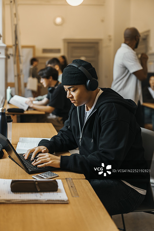 戴着无线耳机的年轻男学生在大学图书馆学习，同时使用笔记本电脑图片素材