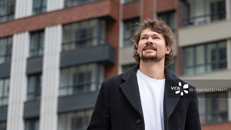白天，一名男子穿着外套，在城市环境中抬头仰望一座现代建筑。图片素材