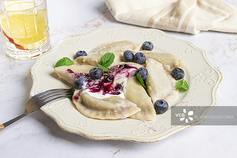 手工制作的甜味饺子，精美地摆放在白盘上，淋上浆果酱，撒上新鲜蓝莓和薄荷叶，准备享用图片素材