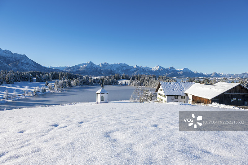 冬季的风景，有农场和教堂，坐落在蓝天下的雪山前，位于德国巴伐利亚州阿尔高地区菲森附近的赫格拉茨里德湖畔。图片素材