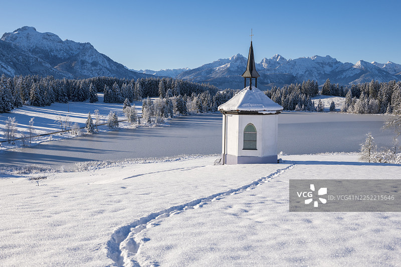 冬天的小教堂，雪地里的脚印，背景是山峦，位于德国巴伐利亚州阿尔高地区福森附近的赫格拉茨里德湖图片素材