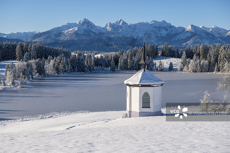 被雪山环绕的冰冻湖岸边的教堂，蓝天下的德国巴伐利亚州阿尔高地区福森市附近的海格拉茨里德湖图片素材