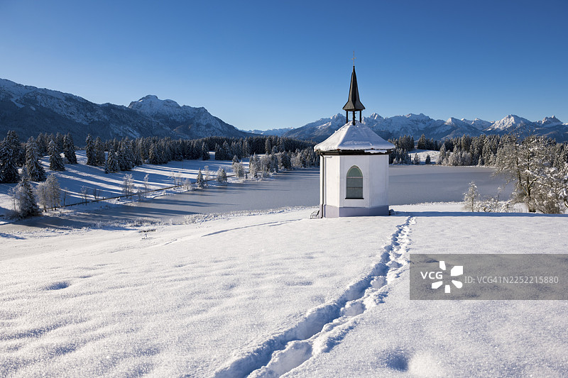 白雪皑皑的雪地里印着脚印的教堂，以及宁静的山景，位于德国巴伐利亚州阿尔高地区福森附近的黑格拉茨里德湖畔。图片素材