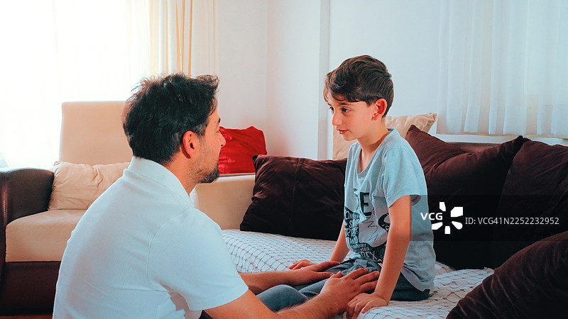 一位父亲和他的年幼的儿子坐在客厅的沙发上，拥抱、交谈并互相开玩笑图片素材