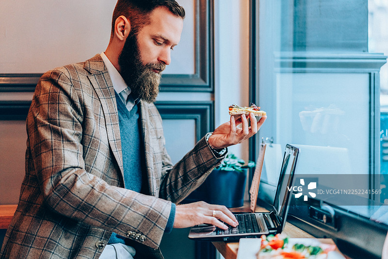 留着胡子的商务人士在咖啡馆一边吃午餐一边用笔记本电脑工作图片素材