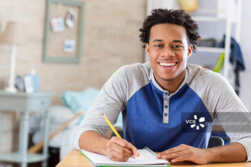 一名年轻的学生正在用铅笔认真地在笔记本上写字，脸上带着灿烂的笑容。图片素材