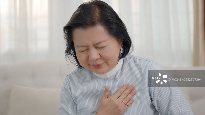 一位年长的妇女独自坐在她宁静、熟悉的家中，双手紧捂着胸口。她脸上因呼吸急促和胸闷而显得痛苦，这可能源于压力或焦虑、惊恐发作、血压异常或神经系统疾病。图片素材