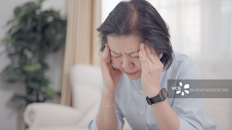 一位年迈的女性坐在家中，感到头晕和剧烈头痛，令她十分难受。她用一只手按住太阳穴，脸上显露出痛苦、紧张和明显的不适。这些症状可能表明她发烧或患有流感，但也可能是其他原因。图片素材
