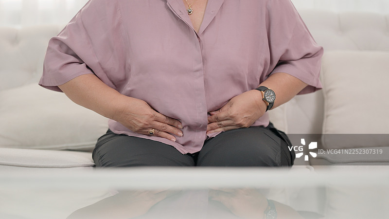 女子独自坐在熟悉而安静的家中，双手紧紧捂着腹部，脸上因腹痛和痉挛而痛苦不堪，这可能由消化系统疾病或慢性健康问题引起。医疗保健与疾病。图片素材
