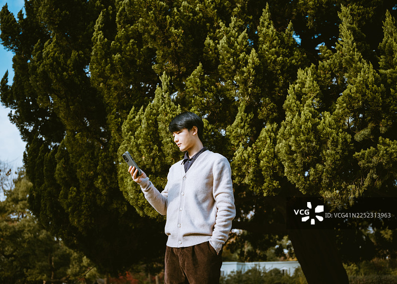 一位年轻的亚洲男子站在一棵绿叶茂密的柏树前，正在使用他的智能手机。图片素材
