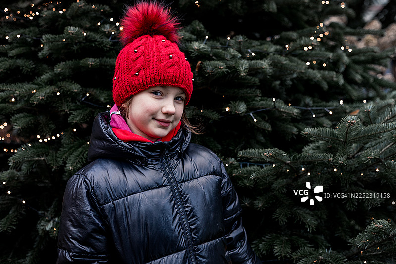 身穿红色针织帽和黑色羽绒服的年轻女孩，置身于圣诞灯和常青树丛中图片素材