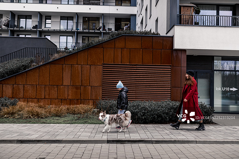 身穿红外套的女子牵着孩子和狗走在现代城市人行道上的城市街道景象图片素材