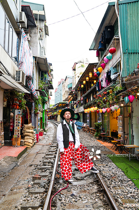 时尚的年轻男子在河内火车街摆出造型，背景是色彩斑斓的建筑、熙熙攘攘的当地生活和舒适的咖啡馆，火车偶尔会穿过狭窄的街道。图片素材