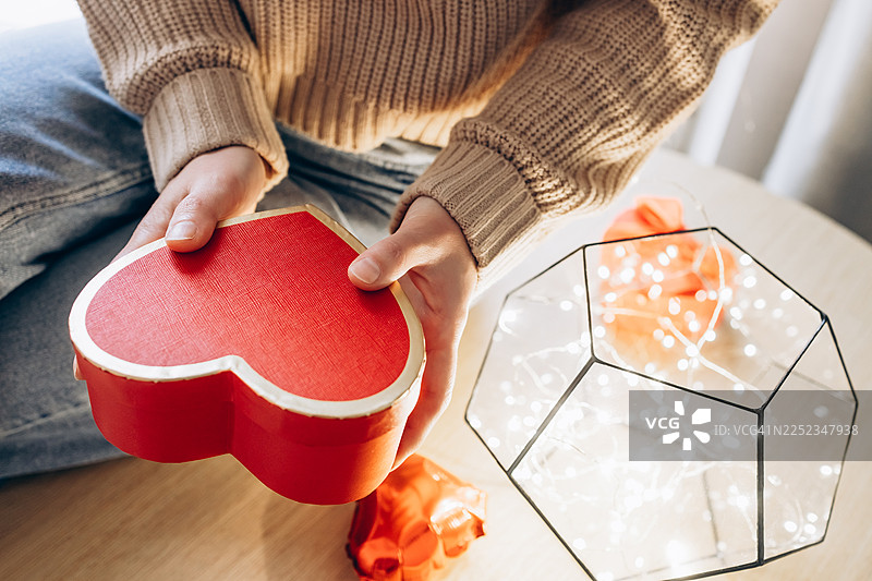 特写：一个人穿着舒适的针织毛衣，在木桌上拿着一个红色的心形礼品盒。图片素材