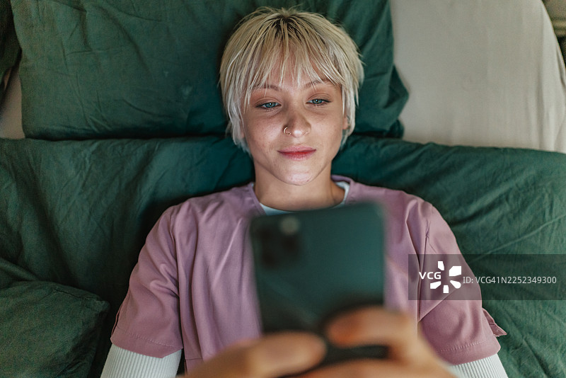 年轻的护士在周末舒适的卧室里，一边使用智能手机，一边享受着悠闲的居家时光。图片素材