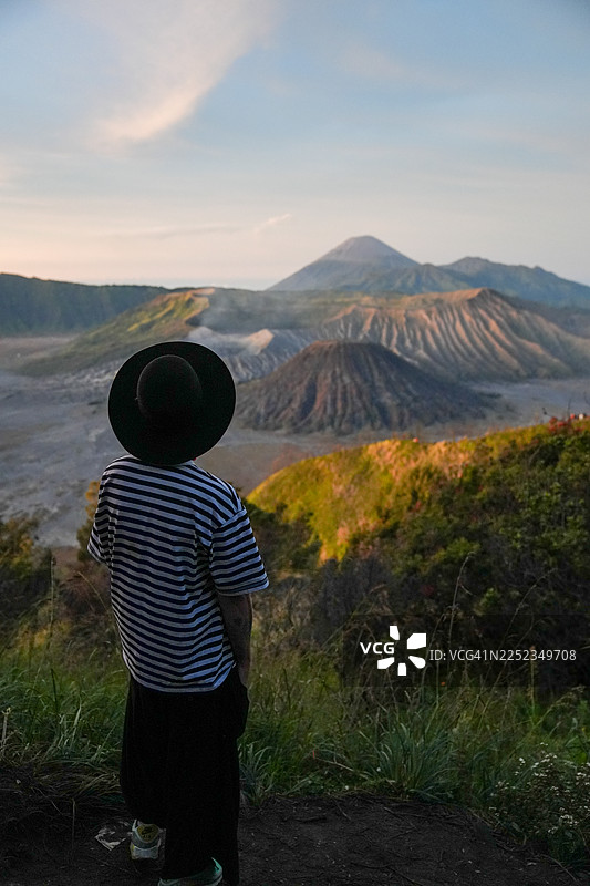 身着时尚服装、头戴帽子的年轻男子站在山脊上，眺望婆罗摩火山的日落景象图片素材