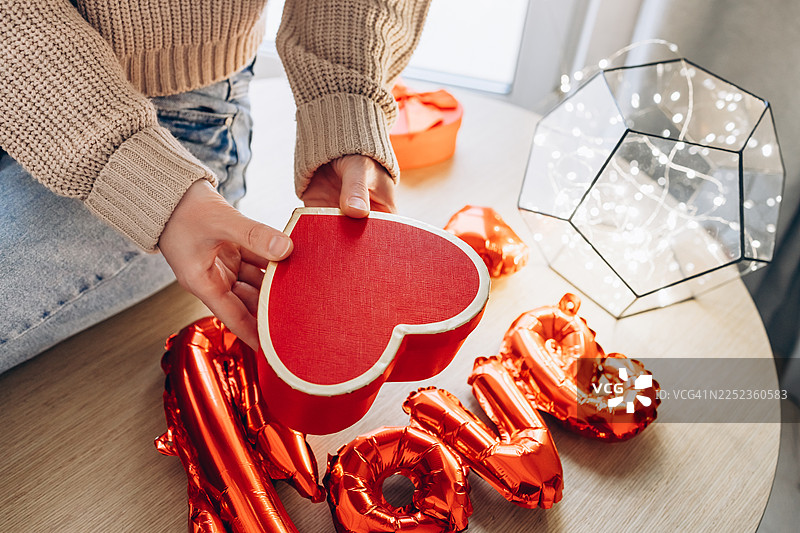 特写：一个人穿着舒适的针织毛衣，在木桌上举着一个红色的心形礼品盒。图片素材