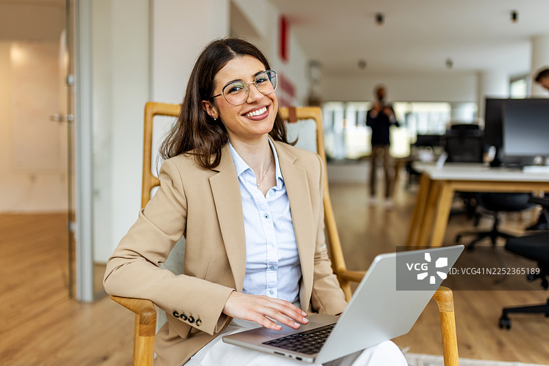 自信的职业女性身着米色西装外套，在现代开放式办公室的笔记本电脑上工作图片素材