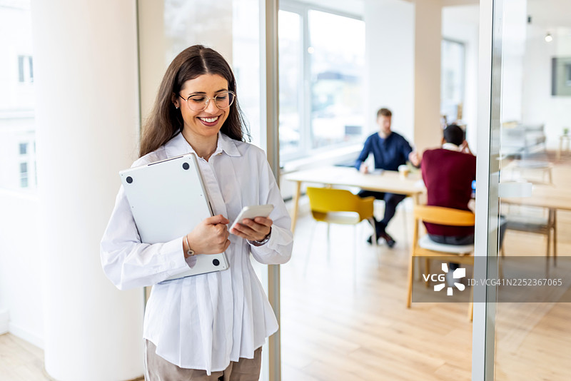 专业女性在现代办公室使用笔记本电脑和手机，同事在背景中协作图片素材