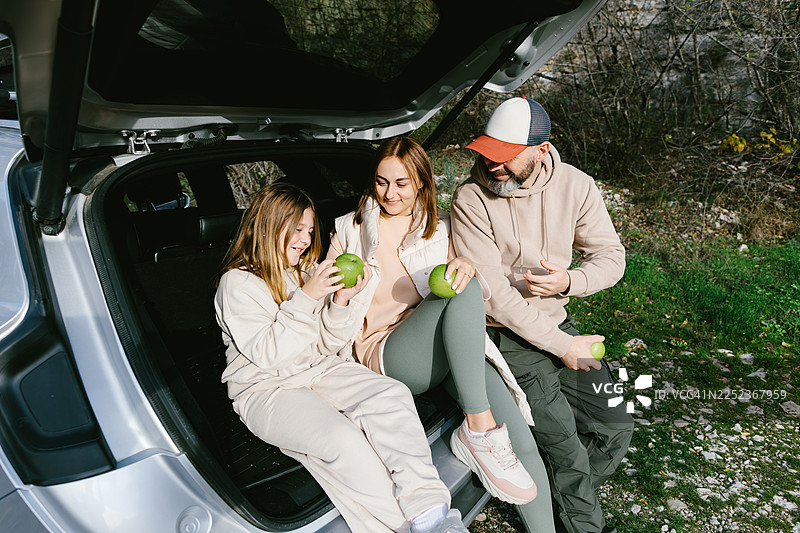 一家人在户外汽车后备箱旁休息，吃着苹果图片素材
