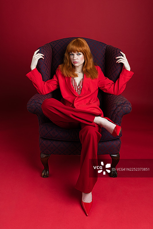 身穿红色套装的自信女性坐在安乐椅上，背景简洁图片素材