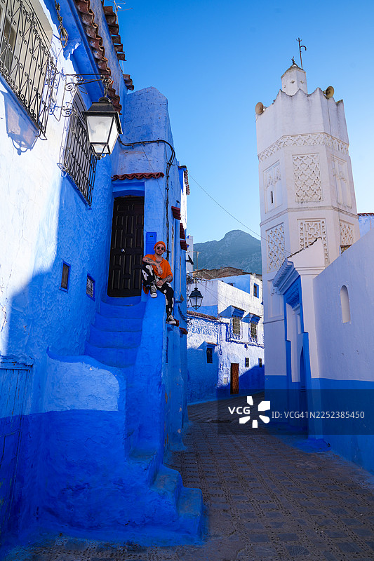 舍夫沙万这座蓝色城市的华丽建筑和街道。一位穿着橙色衣服的时髦男士坐在楼梯上。远处有一座高耸的白色塔楼。图片素材