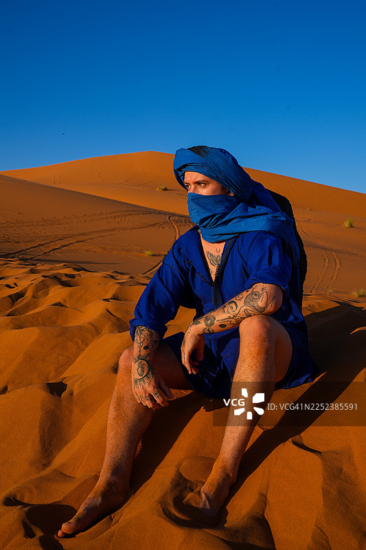 身着蓝色传统服饰的阿拉伯男子独自坐在撒哈拉沙漠的金黄沙丘上图片素材