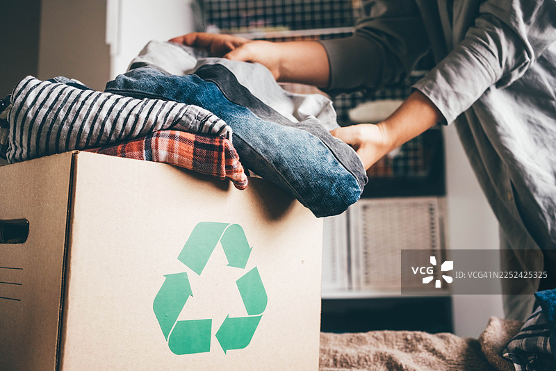 一个人正在打包一个标有回收标志的纸板箱，里面装着各种衣物。这张图片捕捉了可持续生活、道德时尚和环境责任的概念。图片素材
