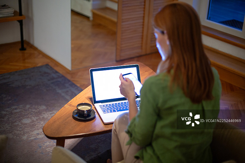 夜晚，一位女士在家中同时使用笔记本电脑和智能手机处理多项事务图片素材