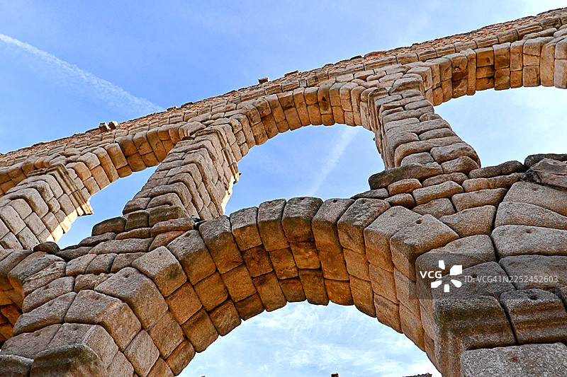 仰视视角拍摄的西班牙塞哥维亚古罗马输水道，背景是湛蓝的天空，展示了其令人印象深刻的石拱和历史工程，塞哥维亚，西班牙图片素材