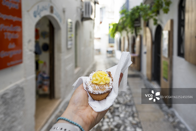 阳光明媚的意大利狭窄小巷中，一只手拿着装有黄色奶油的帕斯蒂乔托糕点，背景是普利亚地区图片素材