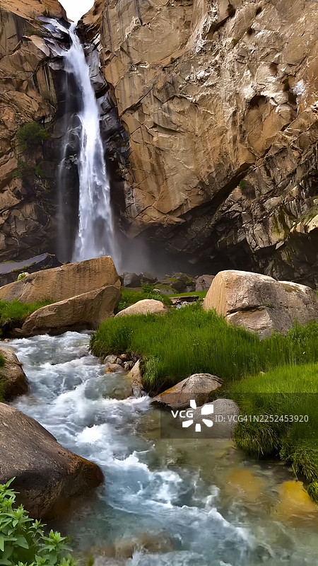 巴基斯坦，雄伟的瀑布从布满岩石的悬崖上倾泻而下，汇入绿草丛生的奔腾河流图片素材