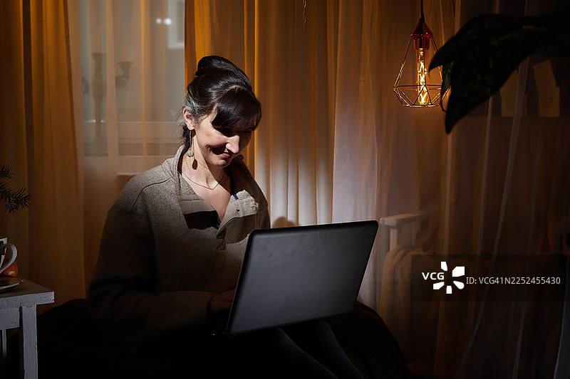 一位女士在家中用笔记本电脑工作，面带微笑，沐浴在温暖的光线下图片素材