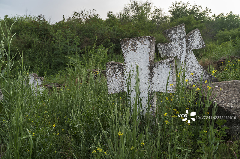 在多云的天空下，杂草丛生的墓地里，古老风化的石十字架掩映在 Tall grass and wildflowers 之中。图片素材