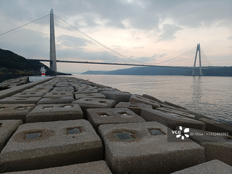 土耳其，一座雄伟的悬索桥横跨宁静的大海，天空布满云彩，前景是混凝土防波堤图片素材
