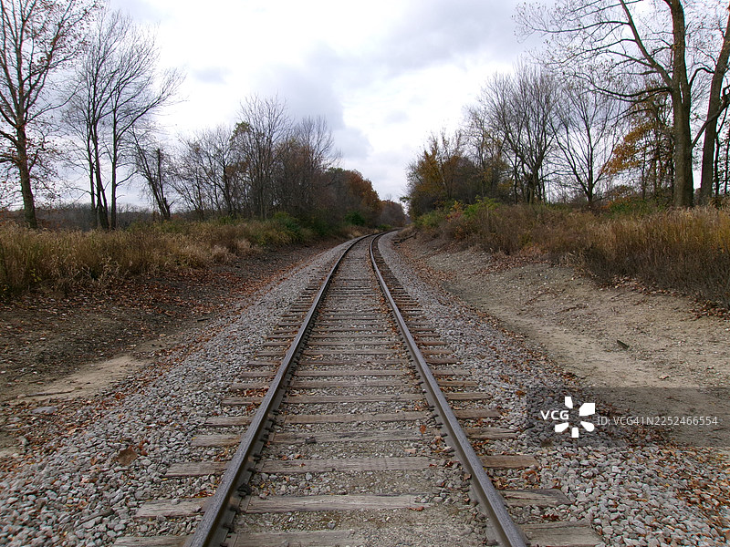 空旷的铁轨蜿蜒穿过树木繁茂的秋季风景，在阴沉的天空下，唤起旅途和孤独的感觉，美国汉密尔顿图片素材