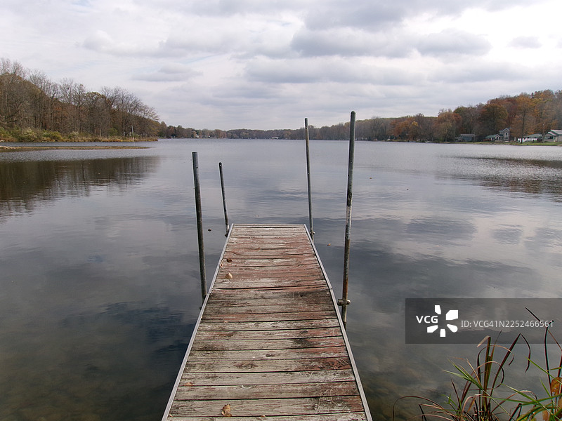 延伸入平静湖泊的木制码头，四周环绕着秋季树木，天空多云，地点：汉密尔顿，美国图片素材