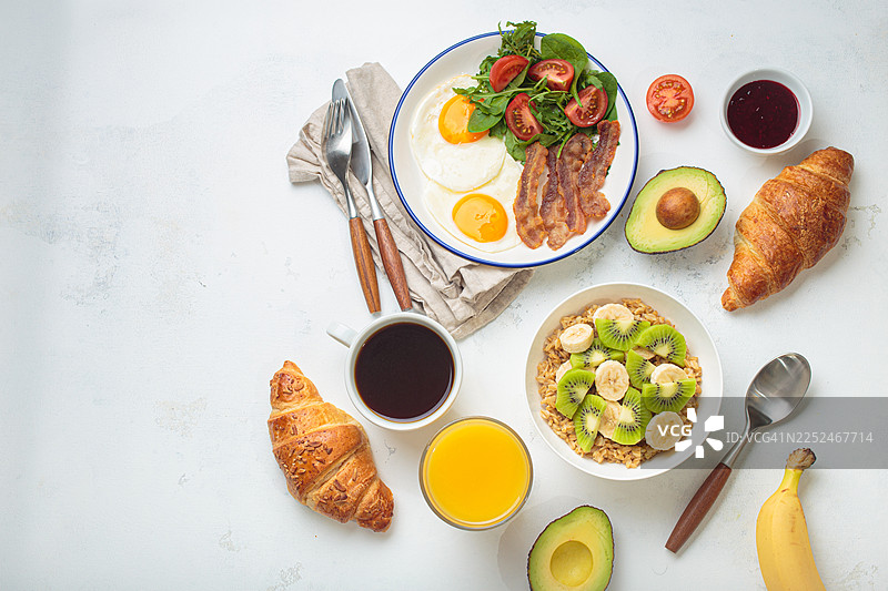 俯视拍摄的各式早餐，包括鸡蛋、培根、羊角面包、水果、咖啡和果汁，摆放在浅色桌子上。图片素材