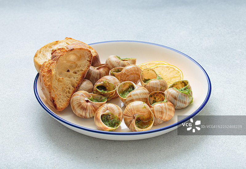白盘上摆放着美味的蒜蓉黄油蜗牛和欧芹，配以新鲜面包和柠檬片，俯视角度拍摄图片素材