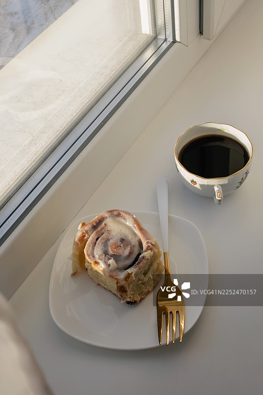 白色背景上，肉桂卷淋着糖霜，旁边放着一杯黑咖啡，透过窗户洒下自然光，俯视视角。图片素材