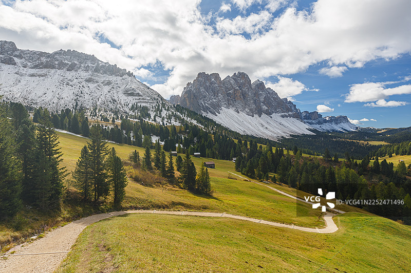 蜿蜒的土路穿过生机勃勃的绿色山谷，远方是白雪皑皑的山峰，天空布满云彩。意大利圣马丁·图尔纳，普埃兹奥德勒自然公园。图片素材