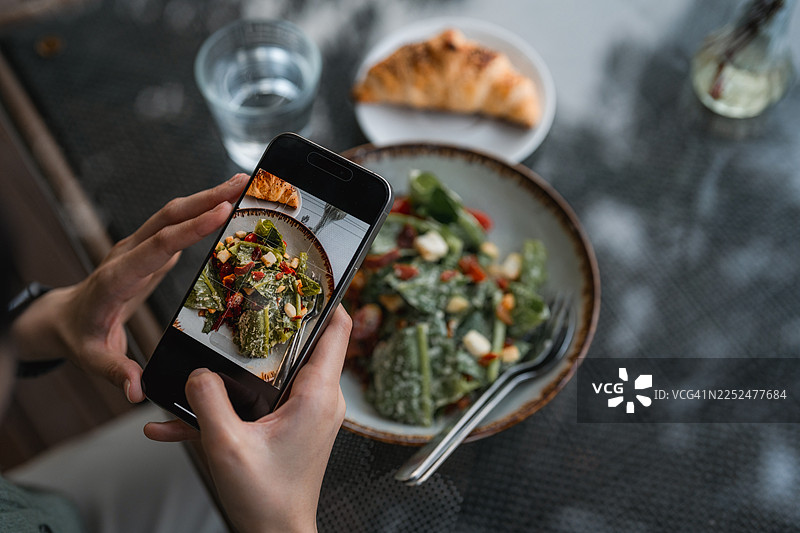 一位快乐的年轻女子在咖啡馆用智能手机为她的食物拍照图片素材