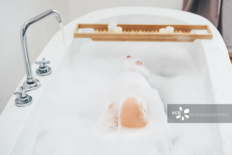 女人光脚泡在现代白色浴缸里，浴缸里溢满了浓密柔软的泡沫。浴缸旁放着一个木质托盘，上面摆放着蜡烛，营造出宁静舒适的居家放松氛围。图片素材