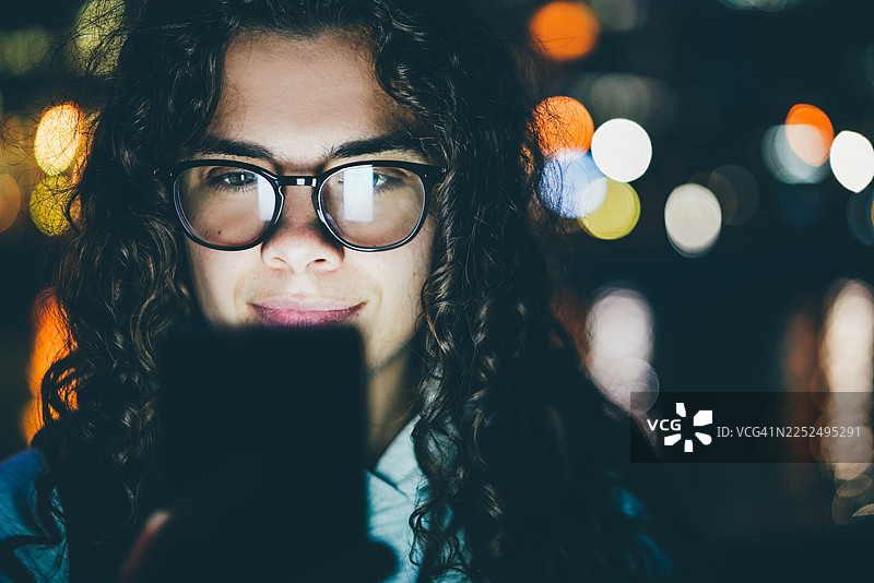 一个留着卷发、戴着眼镜的女人在夜晚看着她的智能手机，模糊的城市灯光背景营造出温馨的都市氛围。图片素材