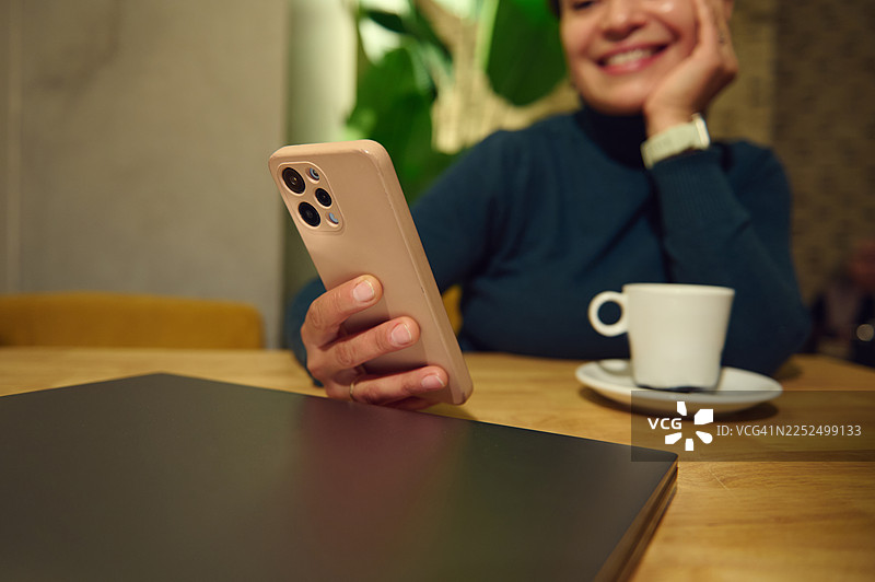 一位女性在舒适的咖啡馆里远程办公，她使用手机和笔记本电脑，面带微笑，身处现代奢华的室内环境。图片素材