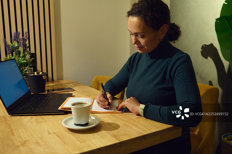中东女性在舒适的咖啡馆靠窗的桌子旁，使用笔记本电脑和记事本进行远程办公图片素材