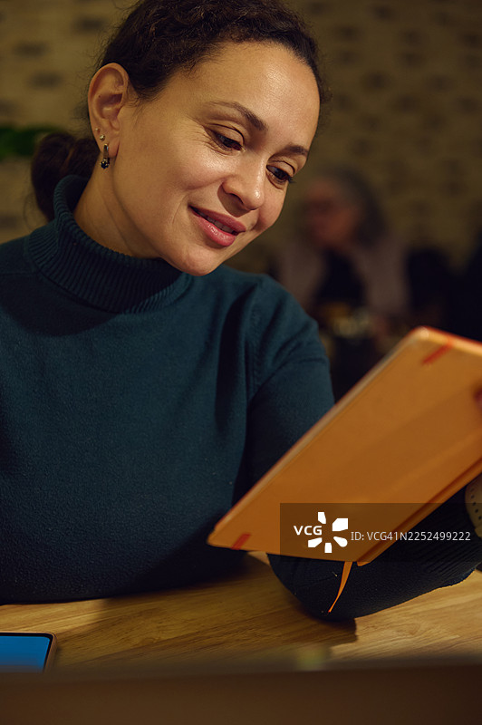 一位专注于工作的远程女员工在舒适的咖啡馆，沐浴在温暖的灯光下，阅读着一本橙色的笔记本。图片素材