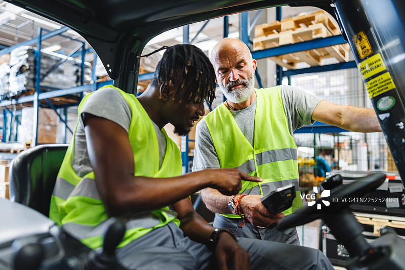 年轻的黑人男性和年长的白人男性在繁忙的仓库中专注地协作处理设备图片素材
