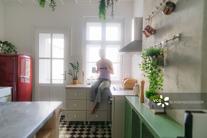 年轻女子在厨房里一边喝茶一边看手机图片素材